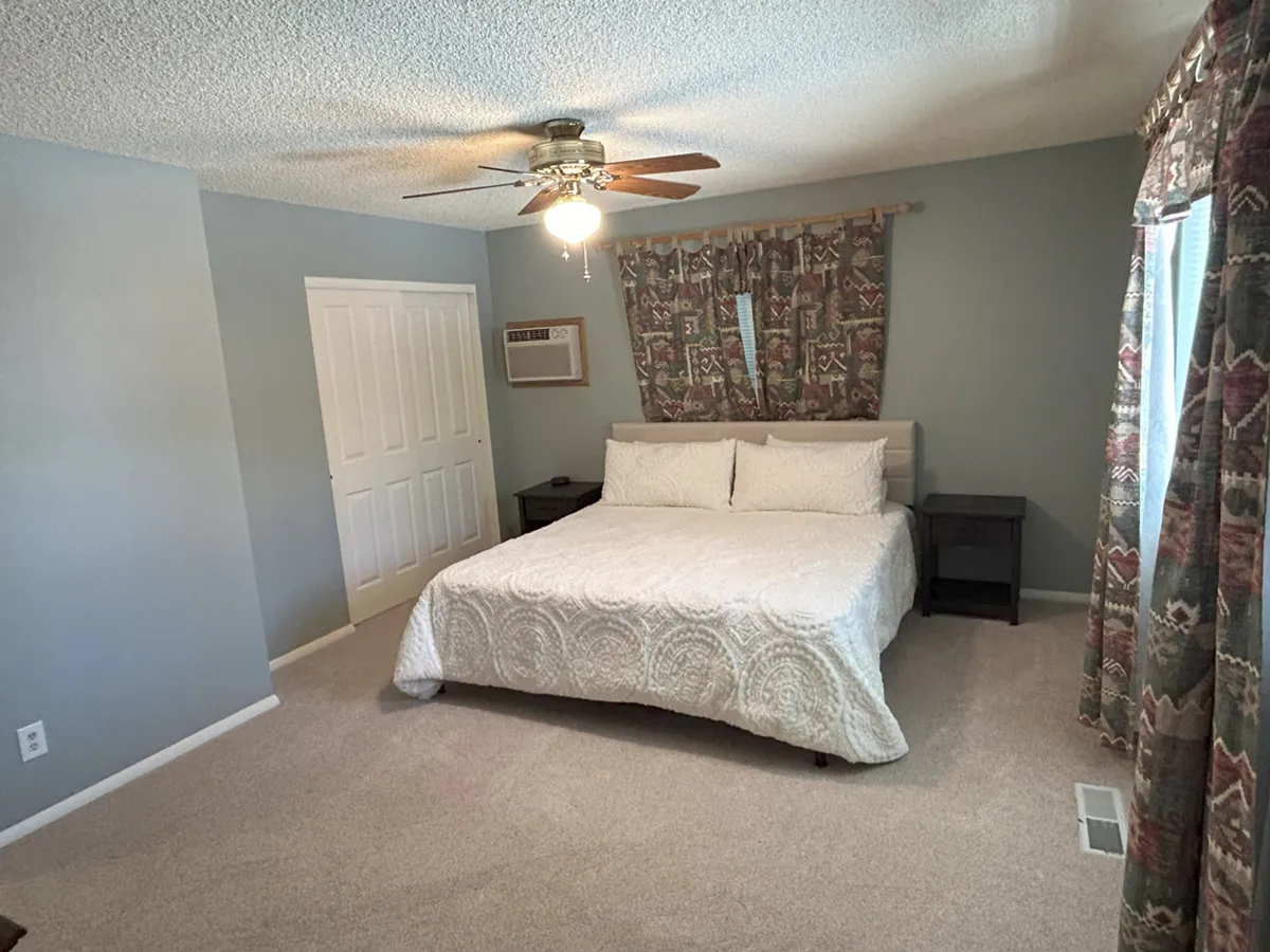 This image features a peaceful bedroom filled with natural light, highlighting a comfortable bed draped in soft linens, curtains framing the window, and a ceiling fan overhead. The mood emphasizes relaxation and the welcoming atmosphere of the home.