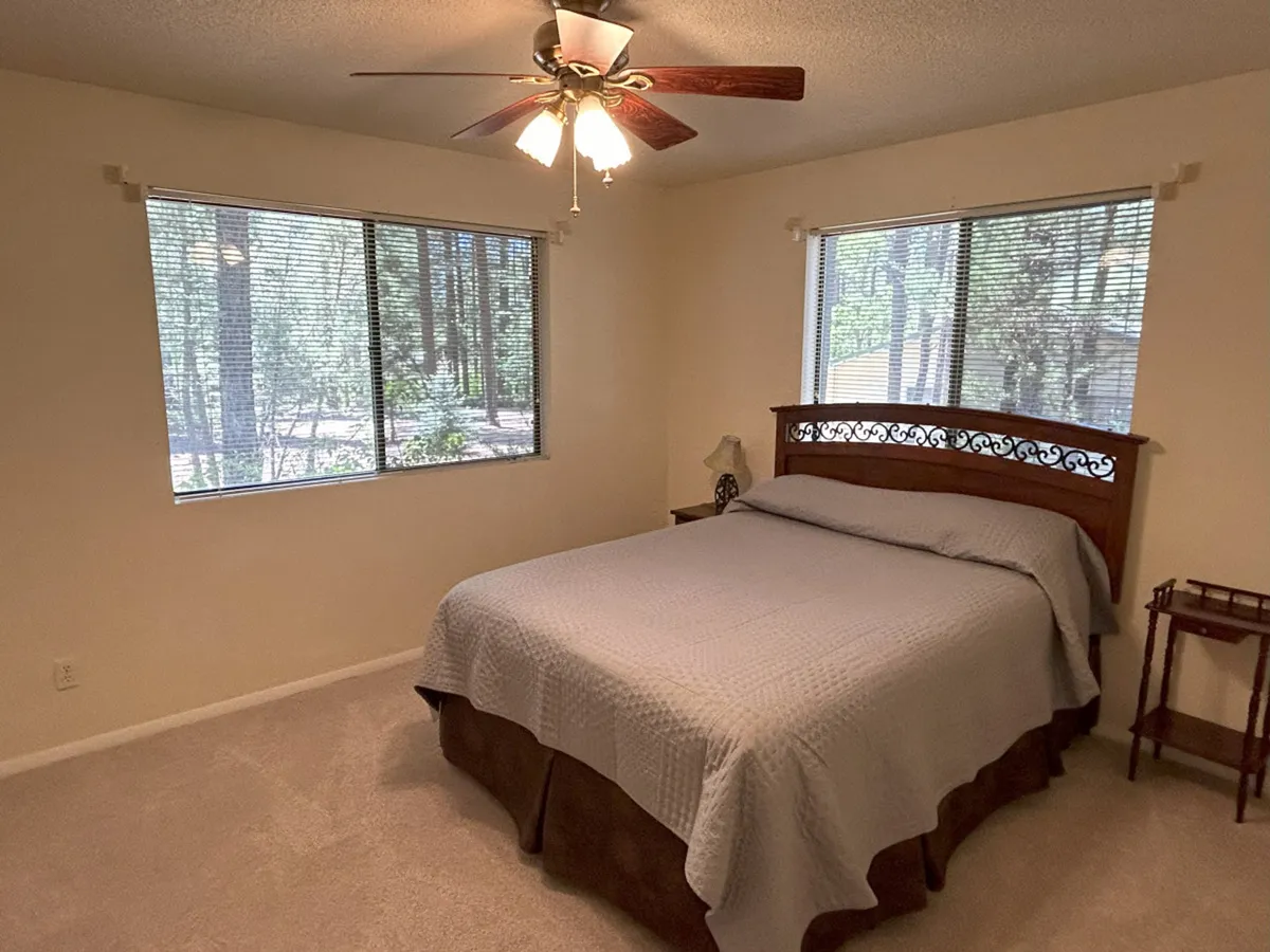 A serene bedroom scene featuring a comfortable bed positioned near large windows that allow natural light to stream in. The setting is tranquil, with tall pine trees visible just outside, creating a seamless transition between indoor comfort and the beauty of nature. Soft furnishings and a nightstand add to the cozy, inviting atmosphere, while a ceiling fan hints at gentle breezes. The overall mood is peaceful and restful, ideal for unwinding.