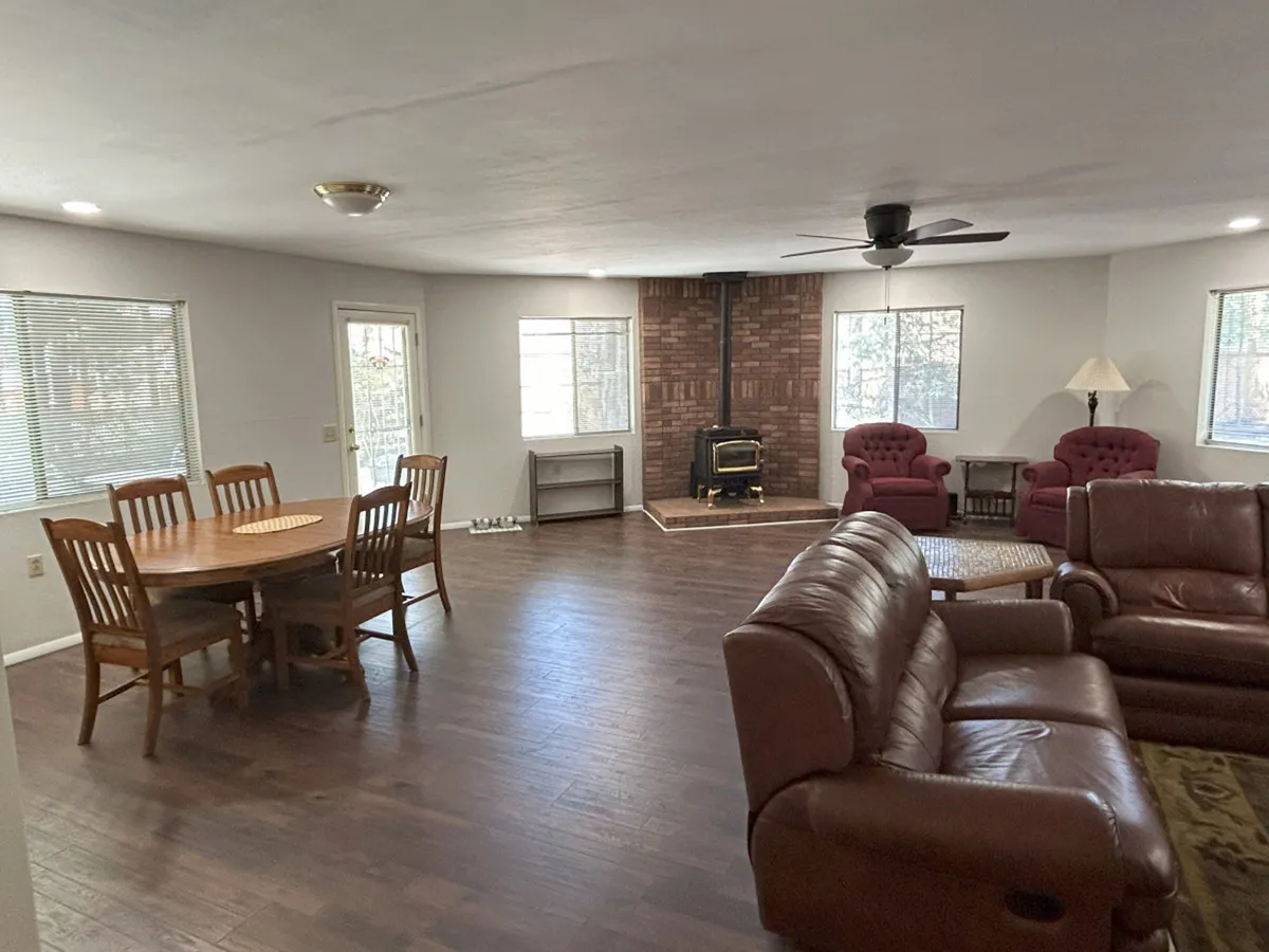 This image portrays a combined dining and living area featuring a wood stove, a spacious dining table, ample comfortable seating, and windows allowing sunlight to brighten the room. The inviting layout is ideal for group activities, cozy evenings by the fireplace, and shared meals, contributing to the overall sense of community.
