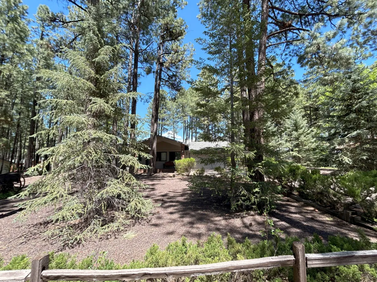 The photo shows a house nestled among mature trees, bordered by a sturdy wooden fence. Lush greenery envelops the property, and the scene conveys direct access to natural surroundings, with trails and wooded views.