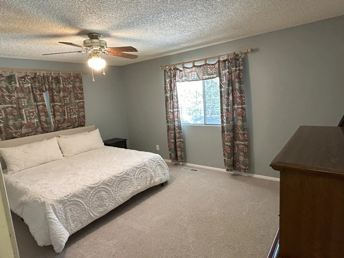 A serene bedroom with a comfortable bed neatly made, sunlight filtering through soft curtains, and a ceiling fan overhead. The window frames leafy green views, creating an inviting space for relaxation and rest.