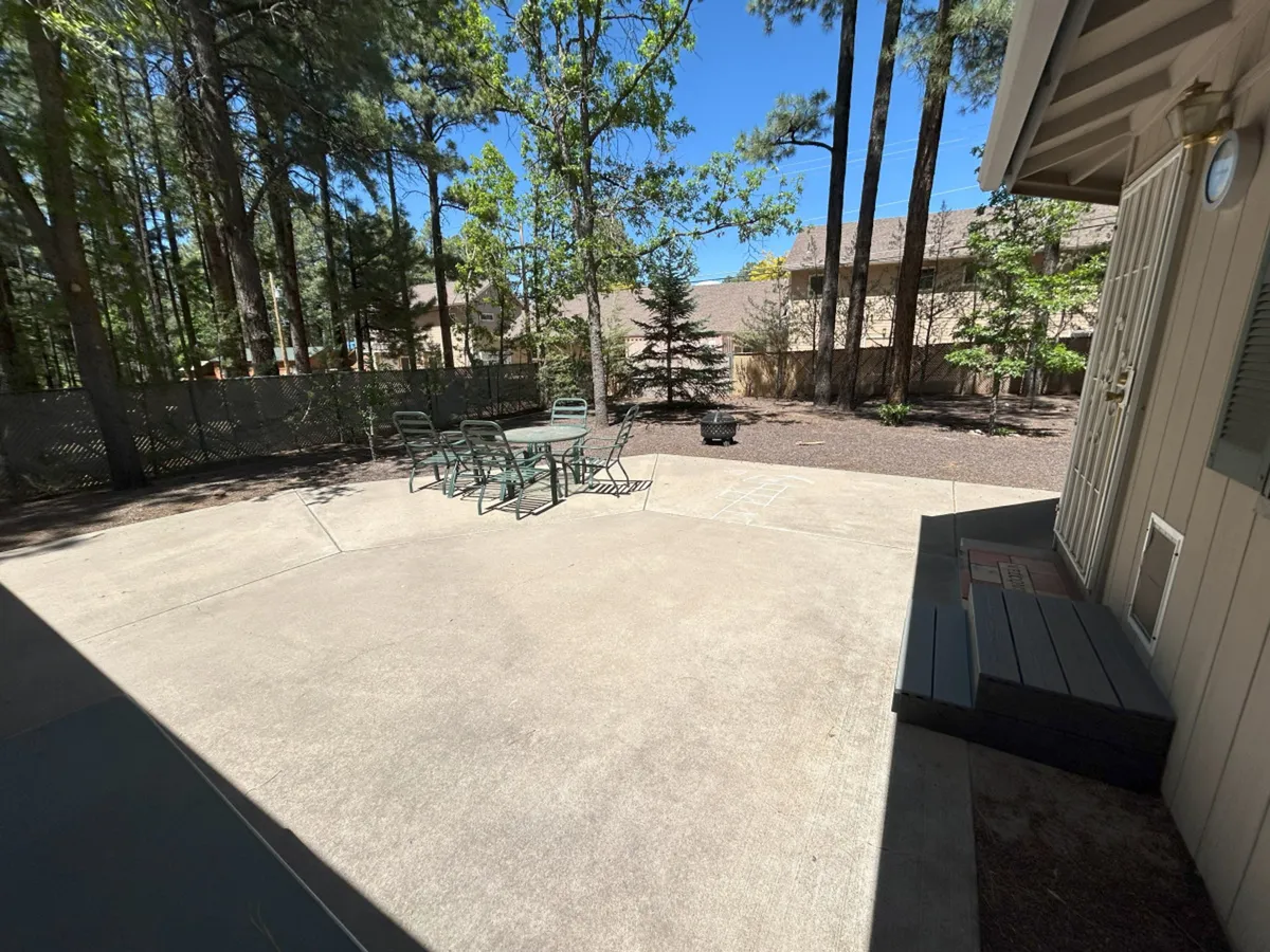 A beautiful outdoor dining area nestled among tall trees, with soft sunlight filtering through the branches. A fire pit sits nearby, surrounded by comfortable chairs, inviting guests to gather and unwind. The natural setting creates a seamless connection between the home and the serene outdoors, perfect for post-adventure relaxation or evening stargazing.